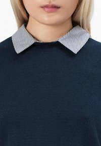 Marineblauer Strickpullover mit einem kontrastierenden gestreiften weißen und blauen Kragenhemd. Weicher Textil mit Rundhalsausschnitt und klassischer Passform.
