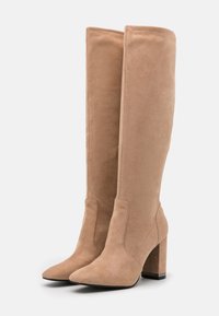 Bottes en daim beige à hauteur de genou avec un bout pointu et un talon épais. Texture lisse, design minimal, avec des détails de couture sur le côté.