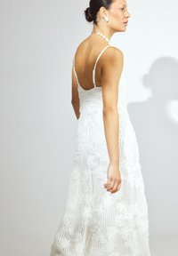Witte maxi-jurk met textuurachtige bloemdetails, spaghetti-bandjes en een open rugontwerp. Glad stof valt tot op de vloer.