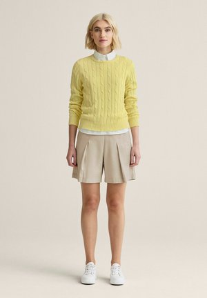 Kvinde med kort blondt hår, iført en gul kabelstrikket sweater, hvid skjorte med krave, beige plisserede shorts og hvide sneakers, stående foran en ensfarvet baggrund.