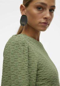 Blusa verde strutturata con un motivo sottile e maniche a sbuffo. Grandi orecchini ovali metallici completano il design.