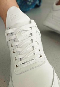 White sneaker with embossed logo, flat laces, metal eyelets, and smooth leather material. Features a sleek, modern design with a rounded toe.