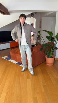 Pilkas striukė su sagomis ir apykakle, dėvima ant balto marškinio. Šviesiai mėlynos džinsai ir sidabriniai sportbačiai. Vidaus interjeras su augalais ir baldais.