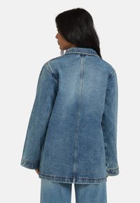 Veste en denim bleu avec une coupe droite, des manches longues et un col pointu. Détails de couture visibles et texture lisse.