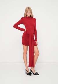 Piros, hosszú ujjú ruha magas nyakkivágással, az egyik oldalon redőzéssel és drapériás részlettel. Fekete, masnival díszített magassarkúval párosítva.