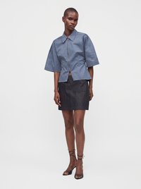 Femme noire portant une chemise bleue zippée à manches courtes, une jupe noire longueur genou avec poches, et des talons ouverts noirs avec brides à la cheville.