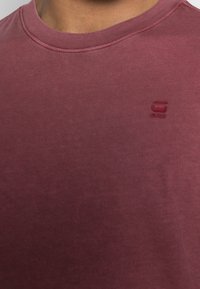 Close-up van een persoon die een kastanjebruin T-shirt met ronde hals draagt, met een klein reliëflogo op de linker borst.