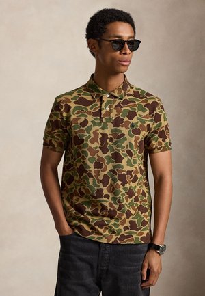 Homme portant un polo camouflage marron et vert, des lunettes de soleil noires, une montre noire et un pantalon foncé, debout avec une main dans la poche.