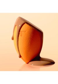 Une éponge de maquillage orange debout avec un liquide marron clair qui coule sur le dessus et s'accumule à sa base sur une surface beige.