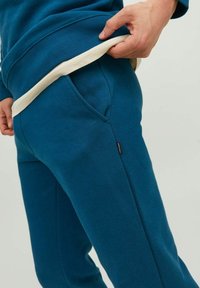 Jogging in teal con una texture liscia, tasche laterali e una vita in crema a contrasto, presenta un piccolo etichetta del marchio sul lato.