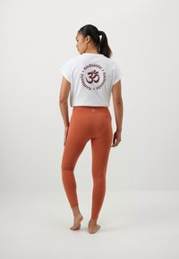 Bílý tričko s maroon symbolem "OM" a textem "namaste" na zadní straně; spárováno s přiléhavými, jednobarevnými oranžovými legínami.
