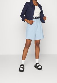 Osoba v námořnickém saku, bílém tílku, světle modrých šortkách s vysokým pasem, bílých ponožkách a černých sandálech s přezkami, stojící na jednobarevném pozadí.