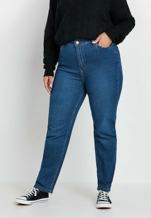 Person iført blå højtaljede jeans med en sort kabelstrikket sweater og sort-hvide sneakers, den ene hånd i lommen, stående på hvid gulv.