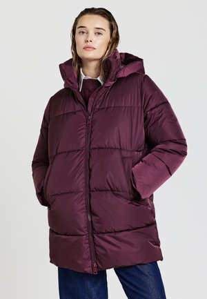 Donna che indossa un giubbotto imbottito viola scuro con un colletto alto, parzialmente aperto su un maglione e una camicia con colletto, con le mani nelle tasche.