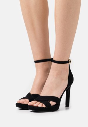 Sandales à talons hauts en daim noir avec une bride à la cheville, bout ouvert et bandes croisées à l'avant. Présente un accent doré sur la bride.