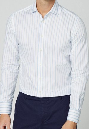 Mann trägt ein weißes Hemd mit hellblauen senkrechten Streifen und navyblaue Hose, steht vor einem schlichten Hintergrund.