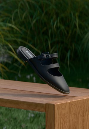 Μαύρο σανδάλι χωρίς λουράκια με δύο ιμάντες και αγκράφα, κρεμασμένο πάνω από ένα ξύλινο παγκάκι με πράσινα φύλλα στο φόντο.