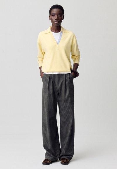Suéter de punto amarillo claro con cuello tipo camisa sobre una camisa blanca; combinado con pantalones de pierna ancha de rayas grises y zapatos con estampado.