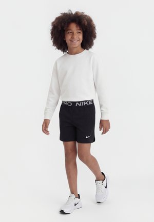 Niño con cabello rizado caminando, vestido con una camiseta blanca de manga larga, pantalones cortos negros de Nike y zapatillas blancas de Nike sobre un fondo blanco liso.