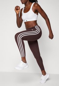 Bílé sportovní podprsenka a hnědé legíny se třemi bílými pruhy po stranách, doplněné bílými sportovními botami. Důraz na aktivní pohyb.