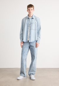 Camicia di denim azzurro chiaro con taschino sul petto, abbinata a jeans azzurri chiari e sneakers bianche, che mostrano una vestibilità lâche e una texture morbida.