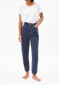 Högmidjade mörka denimjeans med avslappnad passform och lätt avsmalning. Matchas med en löst sittande vit bomullströja. Barfotad.