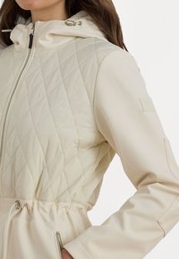 Quilted light beige jacket with a drawstring hood, elastic waist, and zipper pockets; features smooth fabric and subtle branding on the sleeve.