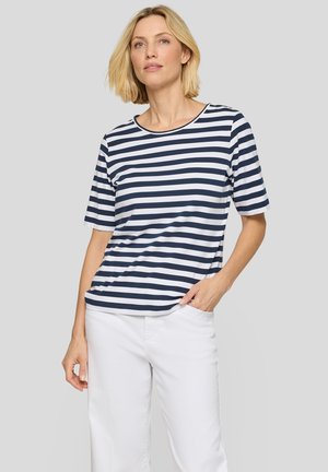 Blonde Frau trägt ein navy-weiß gestreiftes kurzärmeliges Shirt und weiße Hose, steht mit einer Hand in der Tasche.