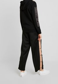 Hoodie noire avec l'inscription "VANS OFF THE WALL" sur la manche, associée à un pantalon noir taille haute avec un détail à bouton-pression rose sur le côté.