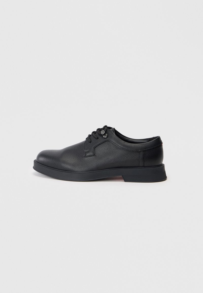 Chaussure habillée en cuir noir avec une surface texturée, bout rond et lacets noirs. Présente un talon bas et une semelle légèrement surélevée.