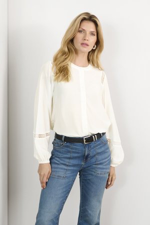 Cremefarvet bluse med knaplukning, blondeaccentuer på ærmerne, parret med højtaljede blå denimjeans og et sort bælte.