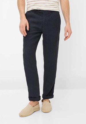 Mand iført sorte bukser med opbuk og beige slip-on sko, stående foran en ensfarvet lys baggrund.