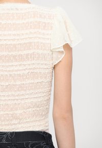 Haut blanc transparent avec manches à volants, présentant une texture froissée et un motif à pois, resserré à la taille.