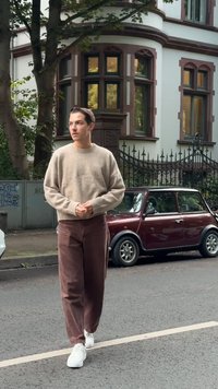 Beżowy sweter z dzianiny z prążkowanymi mankietami, w połączeniu z brązowymi spodniami w szerokim kroju. Białe sneakersy. W tle klasyczny budynek i bordowy samochód.