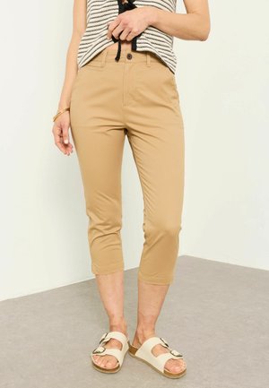Woman wearing beige cropped pants, a striped top, and beige sandals with buckles, standing on a gray floor against a white wall.