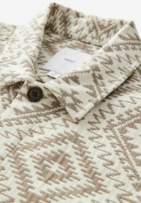 Beige mintás ing, textúrált anyagból, gombos záródással és gallérral; figyelemre méltó, sötétebb tónusú geometrikus minták az anyagon.