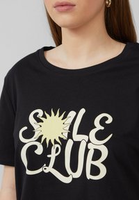 Zwarte katoenen T-shirt met een gele zon-afbeelding en gestileerde tekst "SOLE CLUB" in crème kleur. Ronde halslijn, ontspannen pasvorm.
