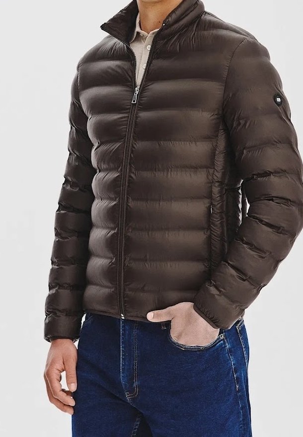 Homme portant une doudoune matelassée marron foncé avec une fermeture éclair frontale, debout avec une main dans la poche d'un jean bleu et l'autre bras détendu.