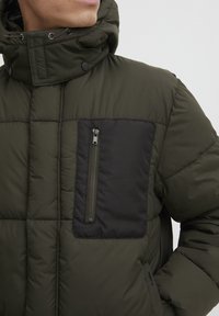 Olīvu zaļš pūkains mētelis ar augstu apkakli, ar melnām zipa kabatām uz krūšu daļas un quilted tekstūru visā virsmā.