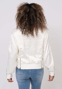 Veste bomber en satin crème avec des manches longues, des poignets côtelés, des détails zippés à l'ourlet et une coupe décontractée. Présente une texture lisse et brillante.