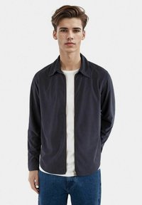 Giacca zip-up blu navale con colletto a punta e maniche lunghe, abbinata a una t-shirt bianca. Tessuto morbido e design a taglio dritto.