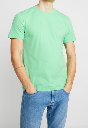 Mand iført en ensfarvet lysegrøn T-shirt med korte ærmer og blå jeans, stående med hænderne i lommerne mod en hvid baggrund.