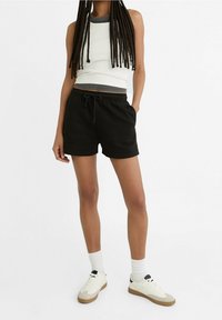 Shorts noirs en coton avec une taille élastique et un cordon de serrage, associés à un débardeur blanc à côtes avec des bordures grises. Baskets blanches et chaussettes.