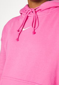 Rosa Nike-hoodie med dragsnodd, mjuk textur och framficka. Har en vit logotyp på övre vänstra bröstet.