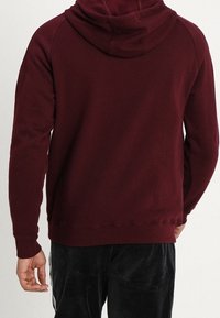 Man som bär en bordeauxfärgad hoodies och svarta byxor, står med ryggen mot kameran.