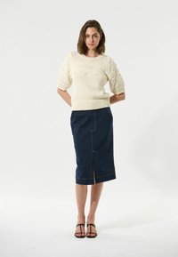 Cremefarvet strikket sweater med prikket tekstur, brede ærmer og ribbet kant. Mørk denim blyantskirt med forslid og kontrastsyninger. Sorte sandaler.