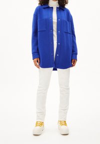 Blå fleecejacka med två bröstfickor och tryckknappar, bärs över en vit polotröja och vita byxor, kombinerat med sneakers med gula snören.