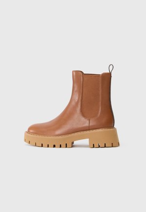 Bottine en cuir marron avec semelle crantée épaisse couleur noisette, panneau latéral élastique et boucle de tirage arrière sur fond blanc uni.