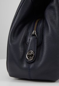 Gros plan sur le coin d'un sac en cuir texturé bleu marine avec une fermeture à tournant argentée et une partie d'une fermeture éclair visible.