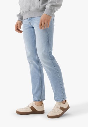 Person trägt hellblaue Jeans, beige Sneaker mit braunem Streifen und graues Sweatshirt, steht vor weißem Hintergrund.